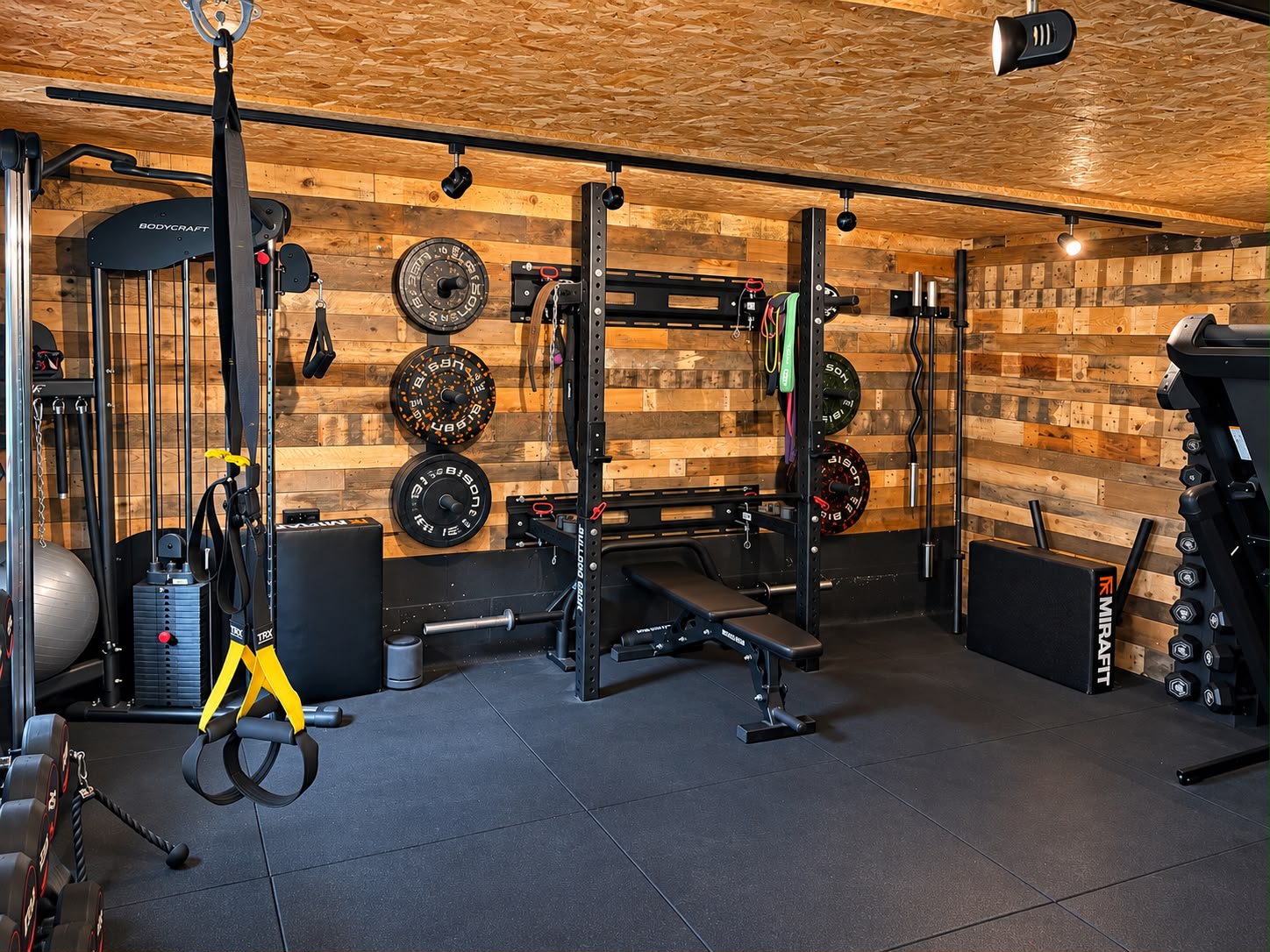 The studio's strength area — squat rack, adjustable bench, bumper plates, cable machine and TRX straps against a reclaimed timber wall